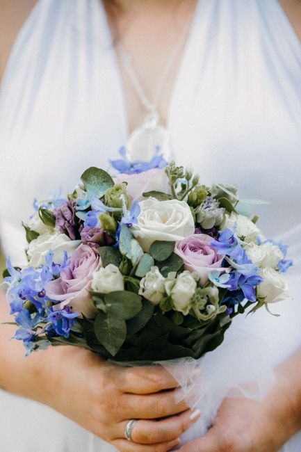 A person dressed in white holding a floral bouquet composed of white roses, light purple lisianthus, and small white spray roses, accented with green eucalyptus leaves and blue delphinium flowers, all