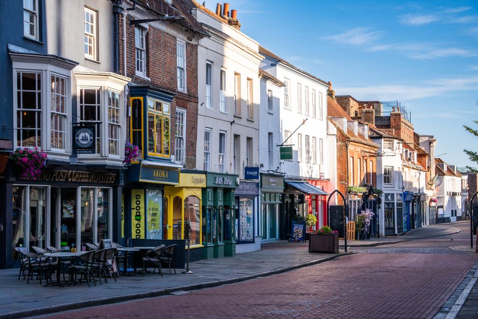 A storefront on a street in Crawley displaying colorful shops with large windows, some decorated with hanging flower baskets. The pavement in front has outdoor seating with black chairs and tables, an